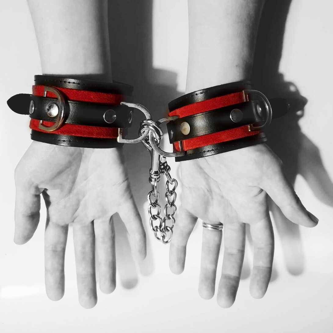 A black and white image of my hands against a white background, wearing leather cuffs with bright red stripes that are attached to each other.